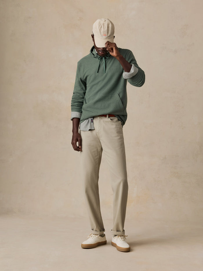 A model stands against a textured beige studio backdrop wearing a sage green long-sleeve polo shirt, light beige trousers, white sneakers, and a cream baseball cap.