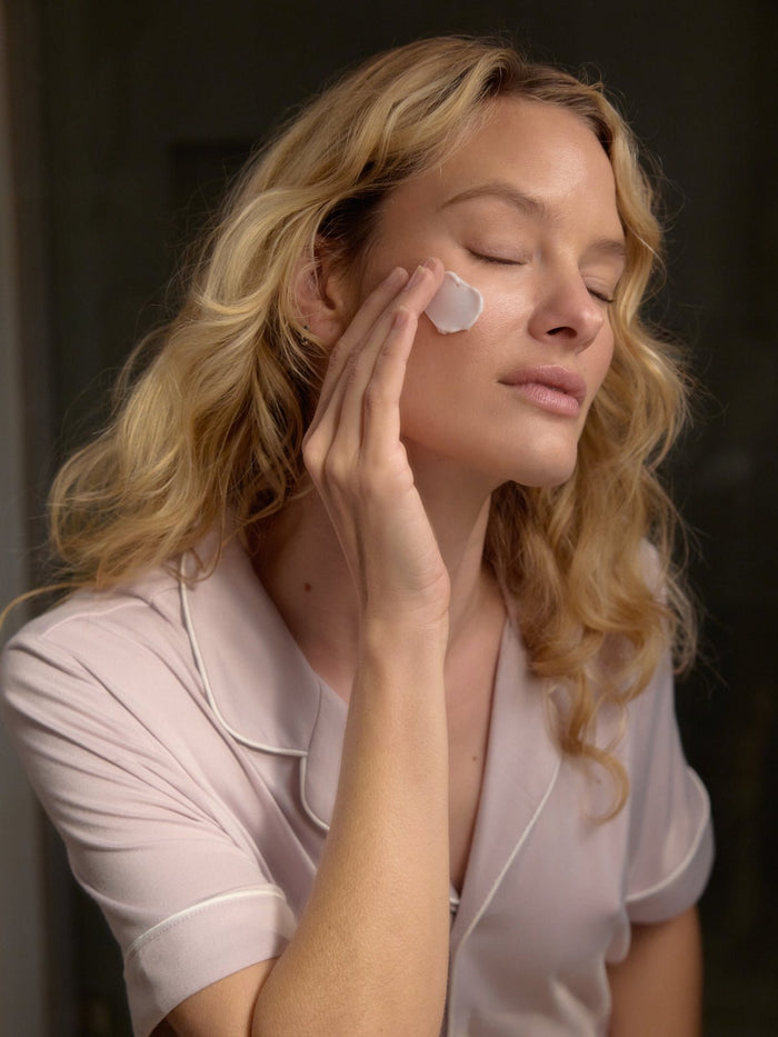 A person with long blonde hair applies a dab of cream to their cheek from the Rest & Reset Bundle by Cozy Earth. Wearing a pastel pink pajama top, their eyes are closed in relaxation. The dimly lit background highlights their calm expression and skincare routine.