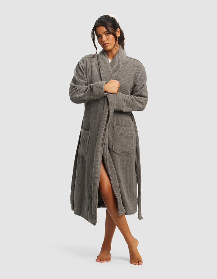A woman with dark hair pulled back stands barefoot against a plain background, wearing Cozy Earth’s Ribbed Terry Bath Robe in gray, featuring front pockets and a waist tie. She looks slightly to the side.|Color:Charcoal