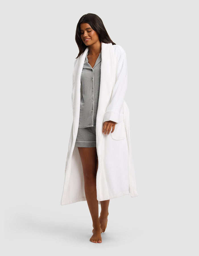 A woman with long dark hair, barefoot and smiling, models the Cozy Earth Ribbed Terry Bath Robe over gray pajama shorts and a button-up top, walking against a plain light gray background.|Color:White