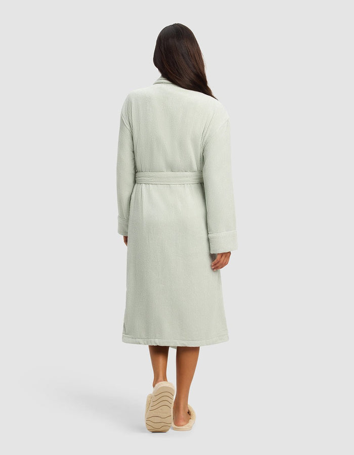 A woman faces away, wearing the Cozy Earth Ribbed Terry Bath Robe in light green with a belt and slippers, set against a plain, light-colored background.|Color:Laurel