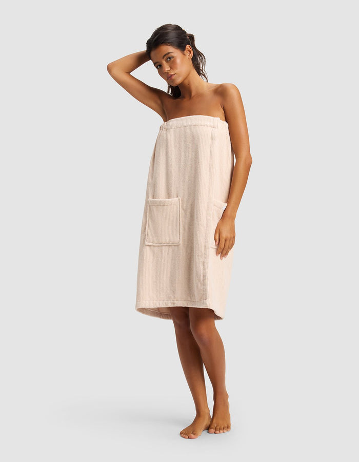 A woman stands barefoot against a plain background, wearing the Cozy Earth Ribbed Terry Bath Wrap with a large pocket, and has one arm raised to touch her head.|Color:Petal