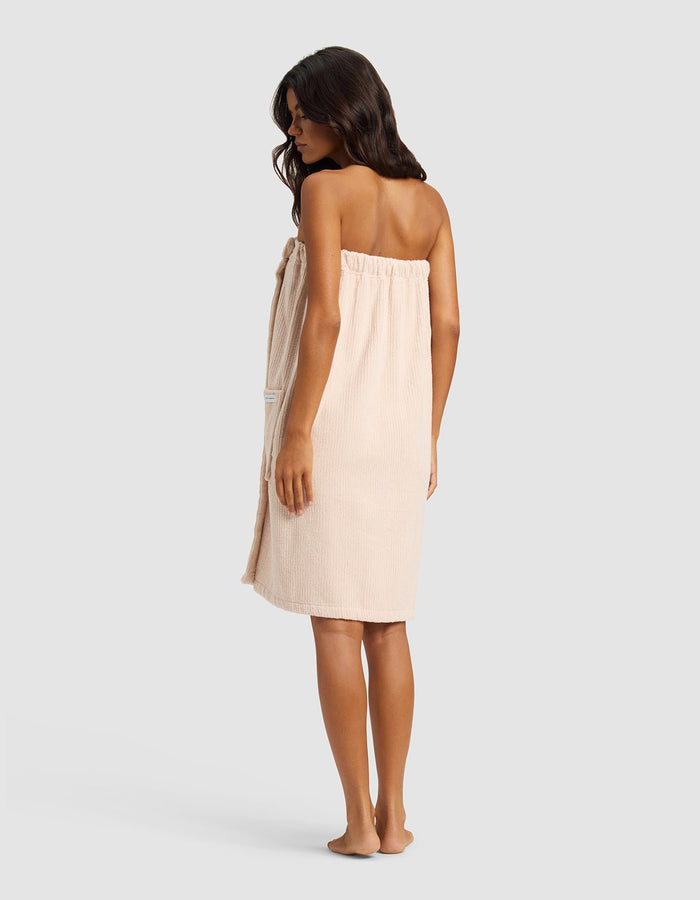 A woman with long dark hair, barefoot and facing slightly away, wears the Cozy Earth Ribbed Terry Bath Wrap in beige against a plain light gray background.|Color:Petal