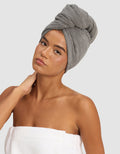 A woman poses against a plain light background, wearing the Cozy Earth Ribbed Terry Hair Towel on her head and a white towel around her body, with one hand gently touching her neck.|Color:Charcoal