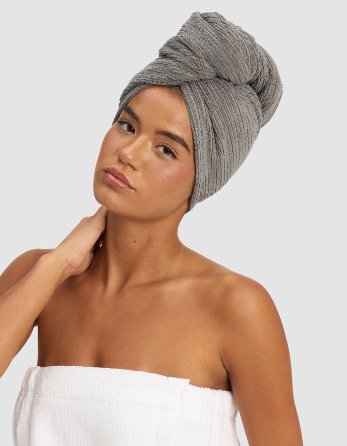 A woman poses against a plain light background, wearing the Cozy Earth Ribbed Terry Hair Towel on her head and a white towel around her body, with one hand gently touching her neck.|Color:Charcoal