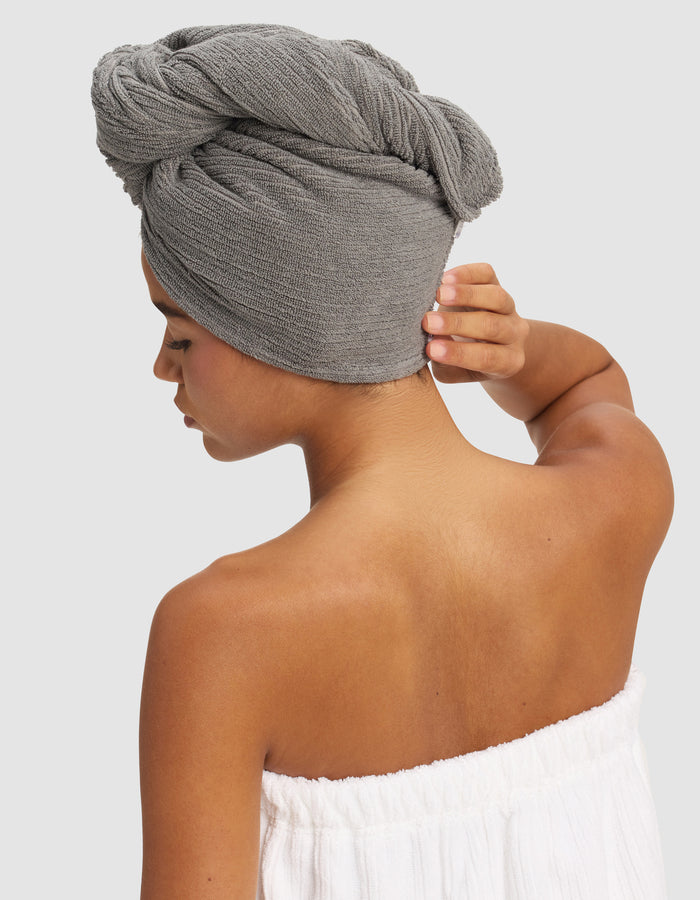 A woman with medium skin tone is seen from the back, wearing a white towel wrap and a Cozy Earth Ribbed Terry Hair Towel in gray, one hand touching the towel on her head, against a plain light background.|Color:Charcoal