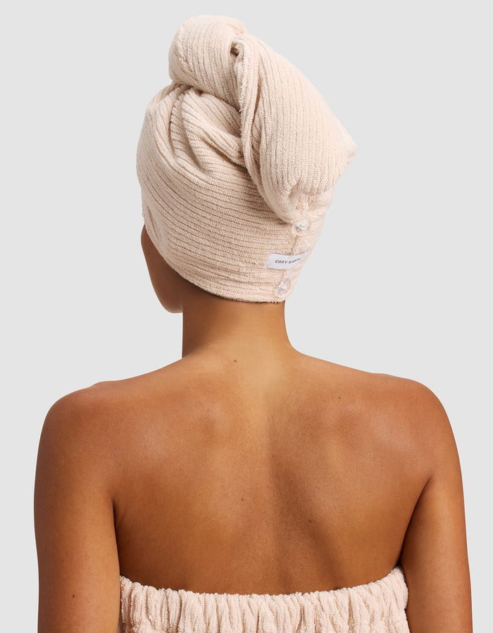 A person with medium brown skin, seen from behind, wears the Cozy Earth Ribbed Terry Hair Towel and a matching strapless towel wrap against a plain light background.|Color:Petal