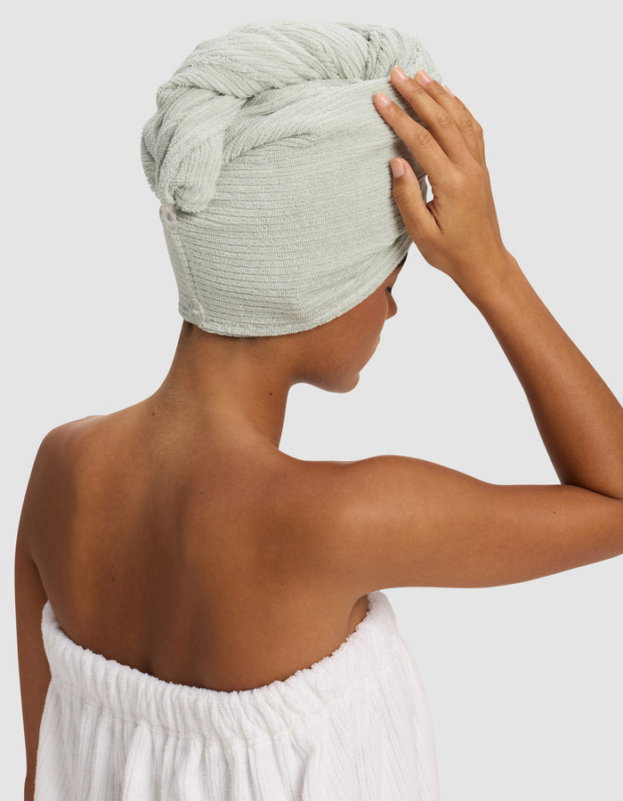 A person with medium brown skin wears a Cozy Earth Ribbed Terry Hair Towel in light green wrapped around their head and a white towel around their body, facing away with one hand touching their head.|Color:Laurel