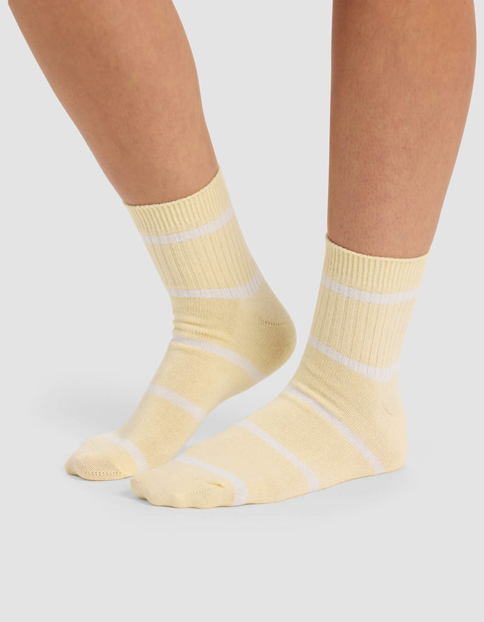 A person wears Cozy Earth Women's Sadie Sock—light yellow crew socks with white stripes—standing on a light gray surface, showing only their lower legs and feet.|Color:Sea Salt/Meringue
