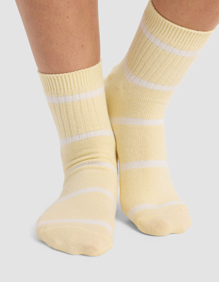 A person stands on a light gray surface wearing Cozy Earth Women's Sadie Socks in light yellow with white horizontal stripes.|Color:Sea Salt/Meringue