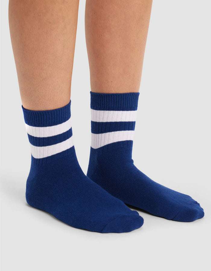 A person wears Cozy Earth's Women's Sadie Sock—blue crew socks with two white stripes near the top—while standing on a light gray background.|Color:Pacific/White