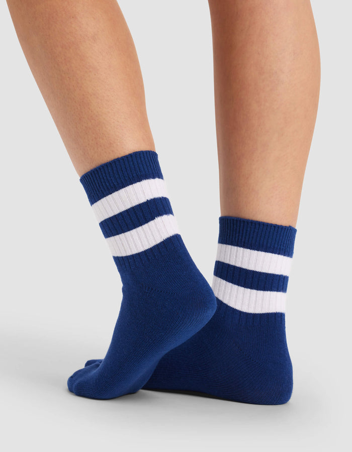A pair of feet wearing Cozy Earth's Women's Sadie Socks in blue with two white stripes near the top, standing on a light gray surface.|Color:Pacific/White