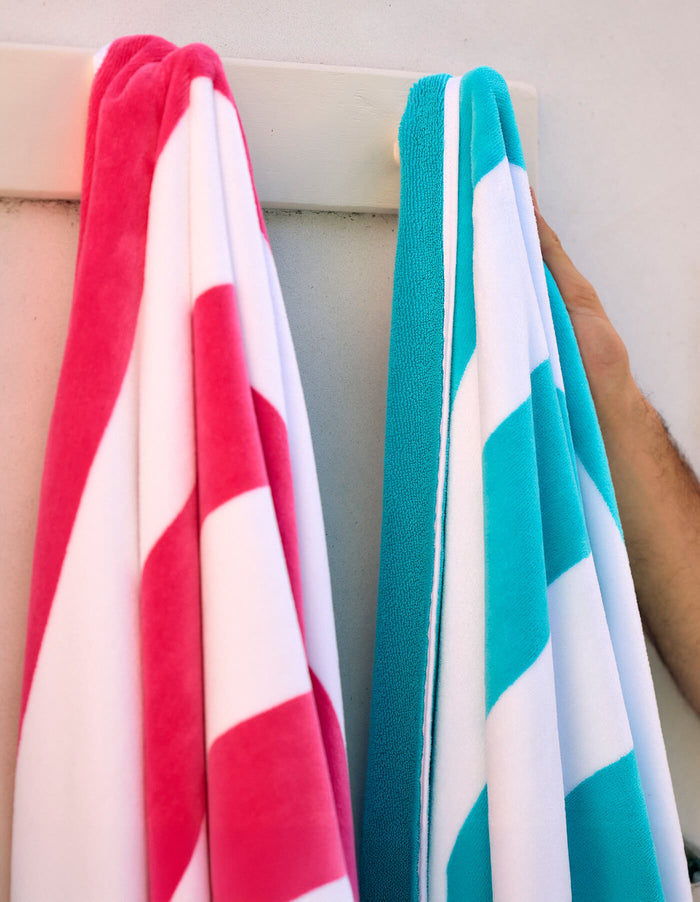 Two Cozy Earth Seaside Resort Towels, one pink and white and the other blue and white, hang on a white rack against a light wall. A person’s arm is holding the blue towel.|Color:Punch Stripe
