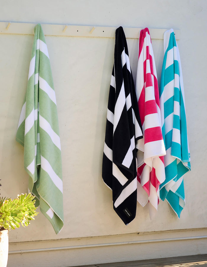 Four Cozy Earth Seaside Resort Towels—in green, black, red, and blue with white stripes—hang side by side on hooks against a light wall. A small succulent plant appears in the lower left corner.|Color:Punch Stripe