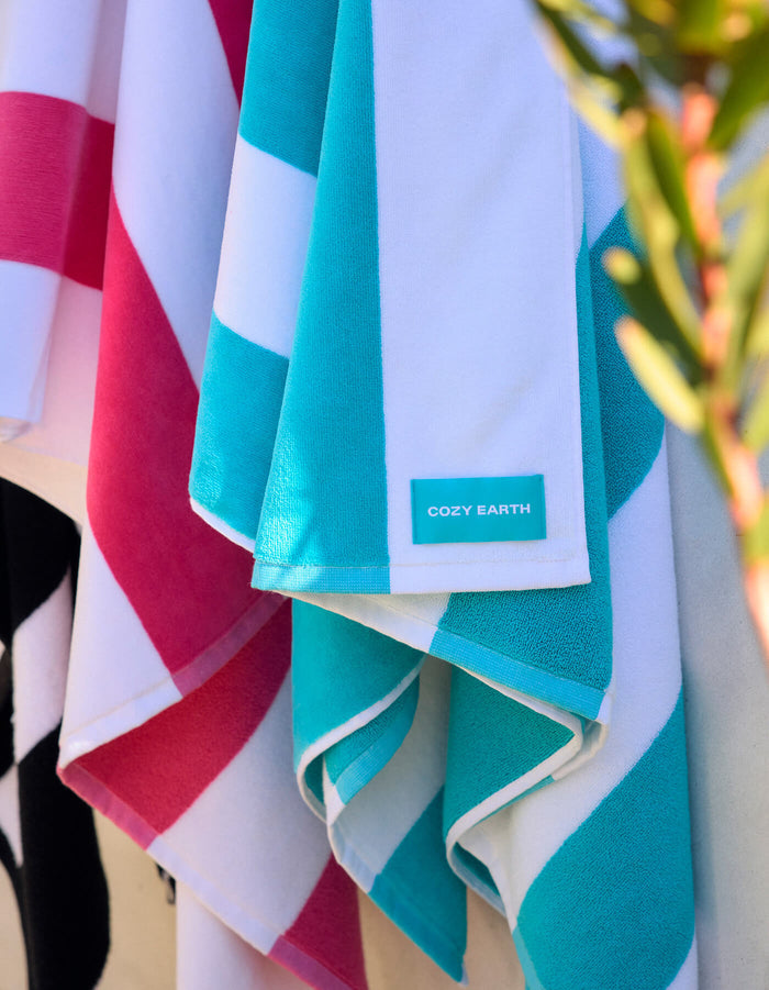 Close-up of Seaside Resort Towels by Cozy Earth hanging side by side in blue, white, and pink stripes. A label reads "Cozy Earth," and a plant branch appears on the right side.|Color:Punch Stripe