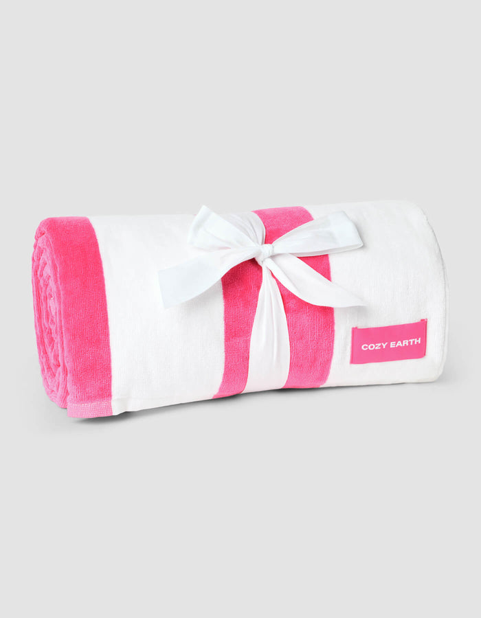 The Seaside Resort Towel by Cozy Earth is a rolled-up white towel with pink stripes, tied with a white ribbon bow and a pink “Cozy Earth” label on the side, displayed against a plain light gray background.|Color:Punch Stripe