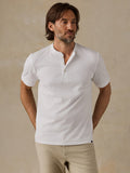 A man wearing a white short-sleeve henley shirt and beige pants, standing against a textured beige wall.