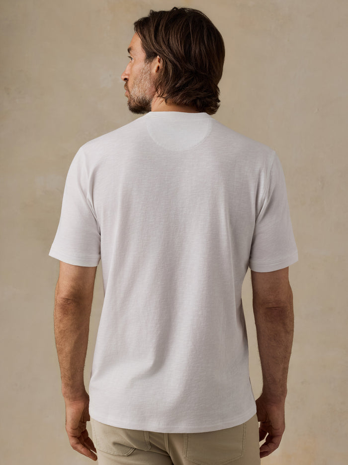 A man viewed from the back wearing a plain white short-sleeve crewneck t-shirt and khaki pants, standing against a textured beige studio wall.