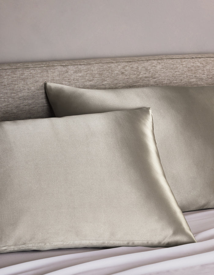 Two Cozy Earth Silk Pillowcases in light gray are arranged on a bed with a light beige upholstered headboard, adding a smooth, shiny, and luxurious touch to the bedding.|Color:Dove Grey