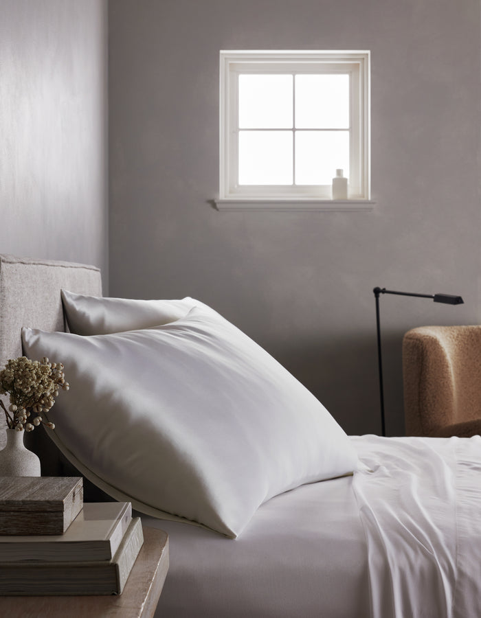A neatly made bed features a Cozy Earth Silk Pillowcase, with white sheets beside a nightstand holding books and dried flowers. Natural light streams in from the window, illuminating a beige armchair and black lamp in the background.|Color:White