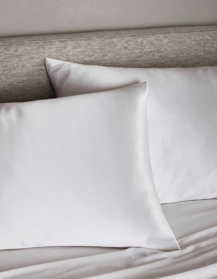 Two Cozy Earth Silk Pillowcases rest on a neatly made bed with a light gray upholstered headboard, creating a clean and minimalistic bedroom atmosphere.|Color:White