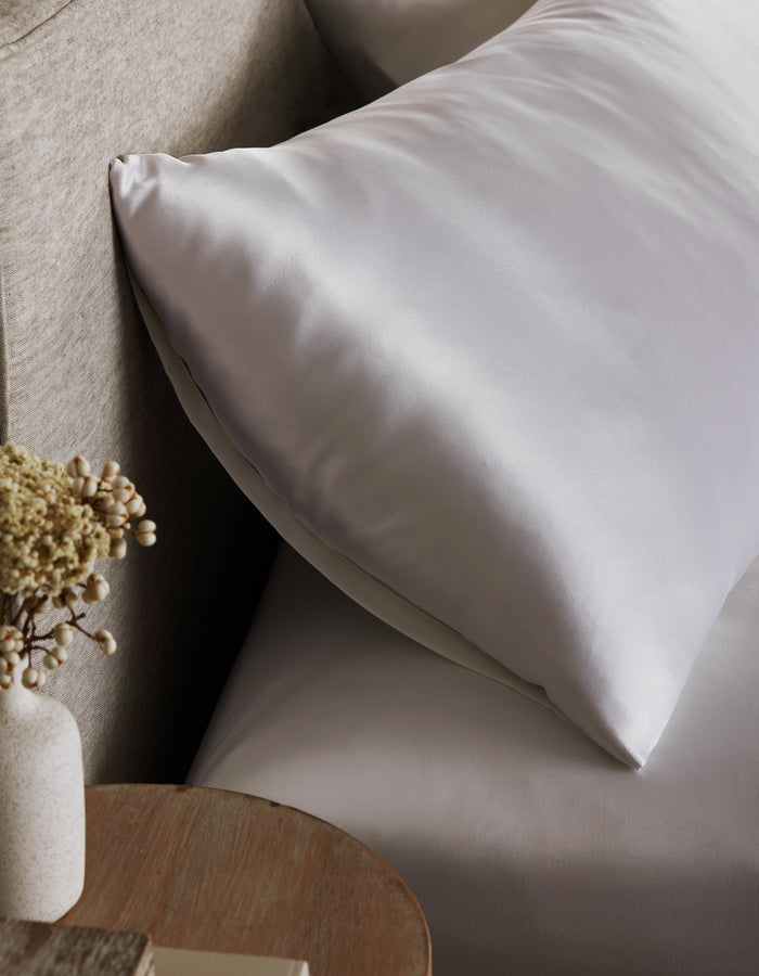 A Cozy Earth Silk Pillowcase sits on a bed with matching sheets by a beige headboard. In the foreground, a small wooden table holds a vase of dried flowers.|Color:White