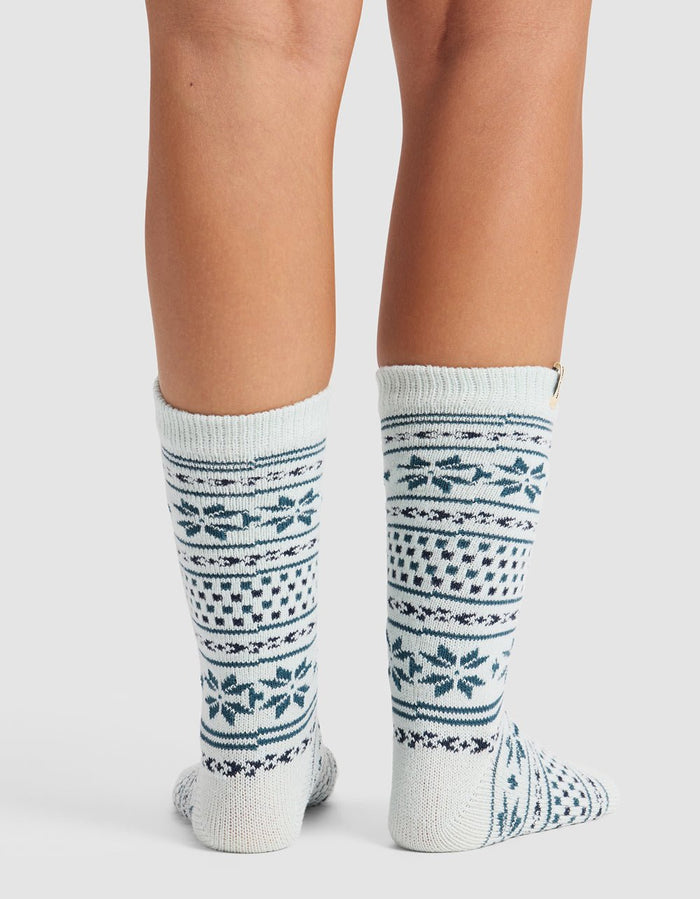 A person wears The Snowflake Plush Lounge Sock by Cozy Earth—knee-high, white knit socks with blue and gray geometric and snowflake patterns—standing with feet together against a plain background.|Color:Mist
