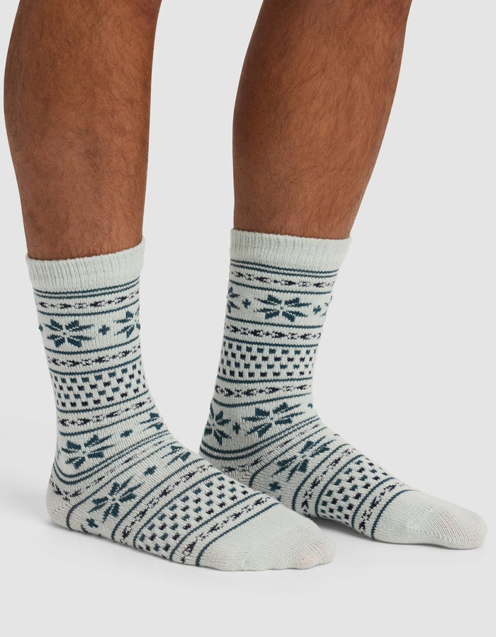 A person wearing Cozy Earth's The Snowflake Plush Lounge Sock—light-colored with dark blue geometric and snowflake patterns—stands on a light surface, showing only their lower legs and feet.|Color:Mist