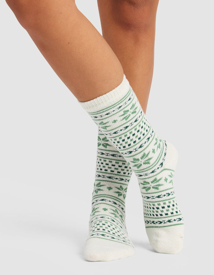 A person models Cozy Earth’s The Snowflake Plush Lounge Sock—white crew socks featuring green geometric and leaf designs—standing with one foot in front of the other against a plain background.|Color:Celadon