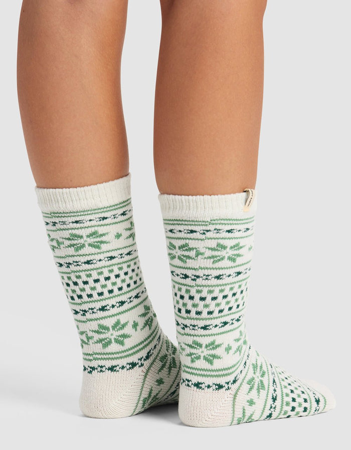 A person wears The Snowflake Plush Lounge Sock by Cozy Earth, featuring cream with green and dark green geometric and leaf designs, standing on a white surface with the back and side of their calves and feet visible.|Color:Celadon