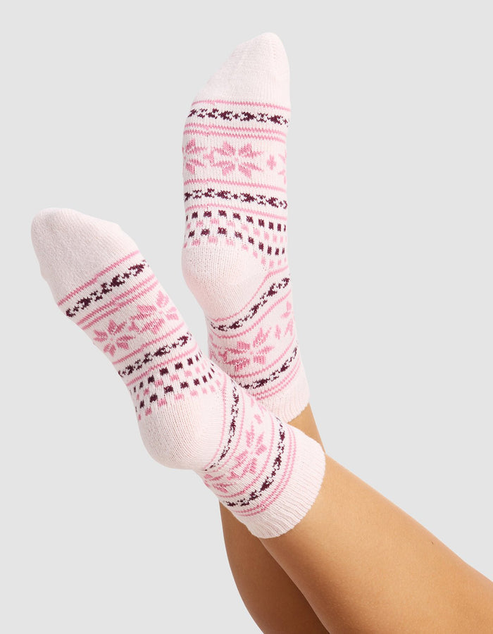 A person wears The Snowflake Plush Modern Crew Sock by Cozy Earth—light pink socks with darker pink and burgundy geometric and floral designs—feet raised against a plain, light gray background.|Color:Ballet