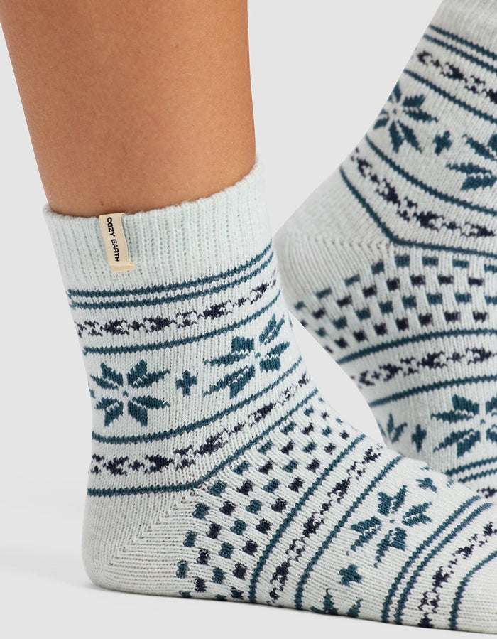 A close-up of feet in The Snowflake Plush Modern Crew Sock by Cozy Earth, featuring light-colored wool with blue geometric and snowflake patterns and a beige cuff tag labeled "Cozy Earth.|Color:Mist
