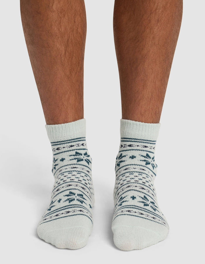A person wearing Cozy Earth's The Snowflake Plush Modern Crew Sock—cream-colored with dark green and blue snowflake and stripe patterns—stands on a white surface.|Color:Mist