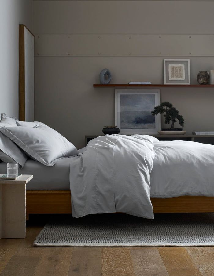 A minimalist bedroom features a neatly made bed with a Cozy Earth Soft-Wash Cotton Duvet Cover in light gray on a wooden frame. Above a dark dresser, art, decor, and a small bonsai are arranged on a floating shelf.|Color:Shore