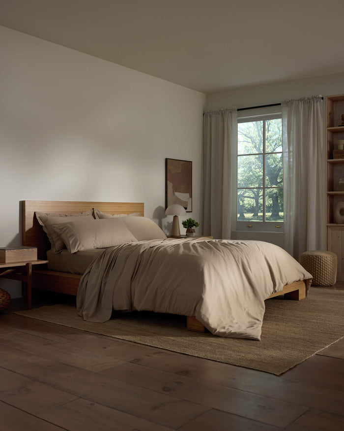 A cozy bedroom featuring a wooden bed frame with Cozy Earth's Soft-Wash Cotton Duvet Cover, a nightstand and lamp, sheer curtains allowing natural light, neutral decor, and wooden flooring.|Color:Sandalwood