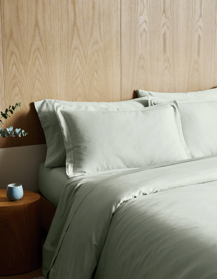 A bed dressed in Cozy Earth's Soft-Wash Cotton Duvet Cover in light green sits beside a round wooden nightstand holding a blue candle and eucalyptus, all set against a light wood-paneled wall.|Color:Laurel