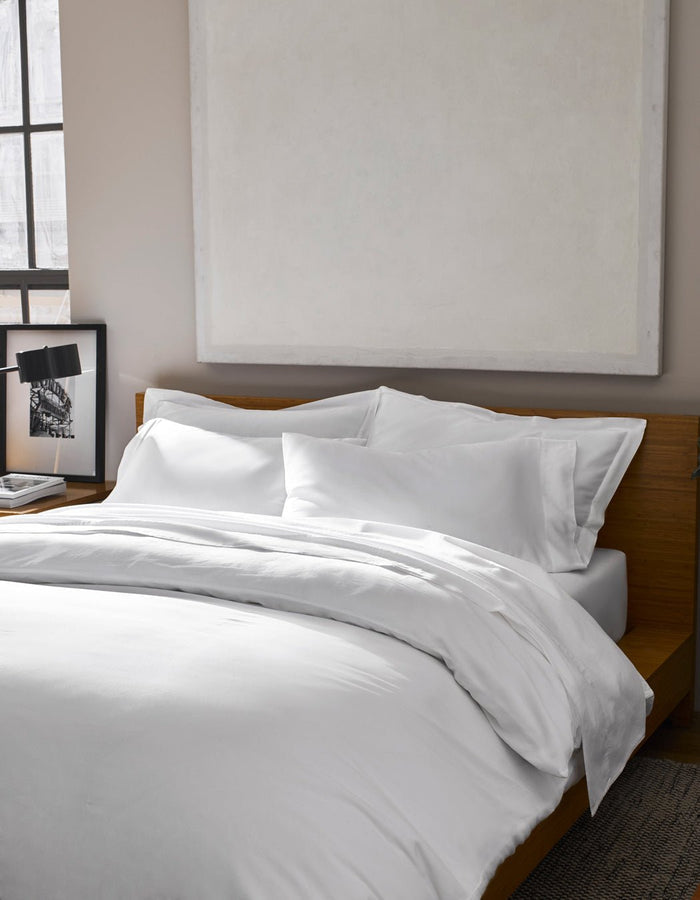 A Soft-Wash Cotton Duvet Cover by Cozy Earth dresses the bed with white sheets against a wooden headboard. Above, a large abstract painting hangs, while a framed photo sits on the bedside table near the window.|Color:White