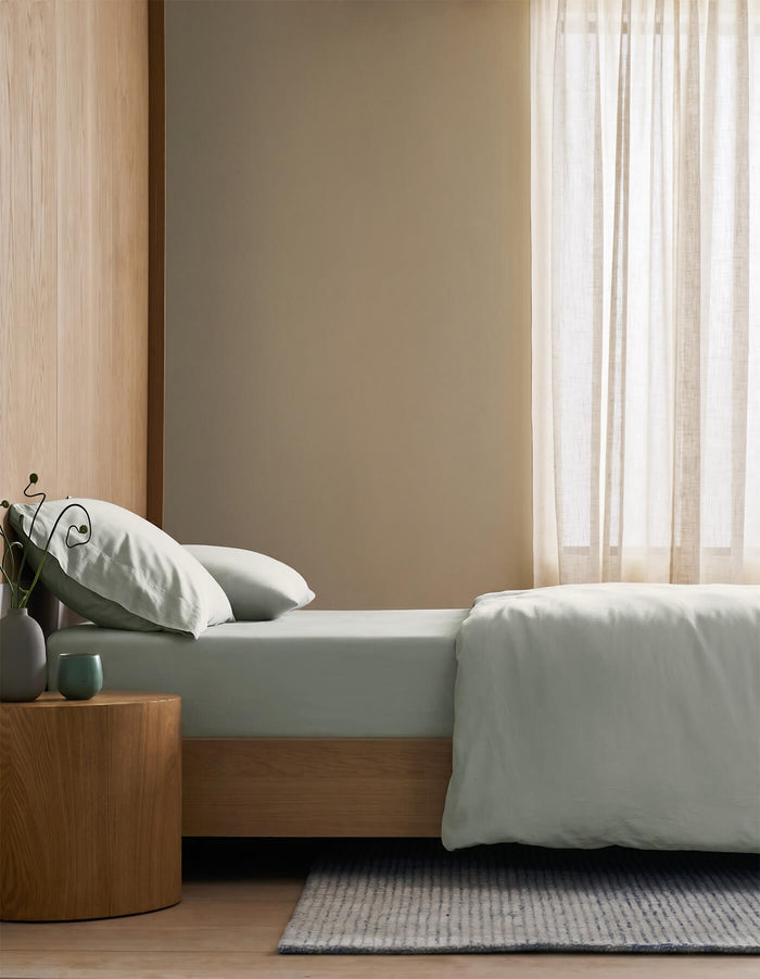 A minimalist bedroom featuring Cozy Earth's Soft-Wash Cotton Fitted Sheet, a light wooden bed, small round wooden side table with a vase, mug and plant, pale gray bedding, light curtains, and a striped rug on the floor.|Color:Laurel