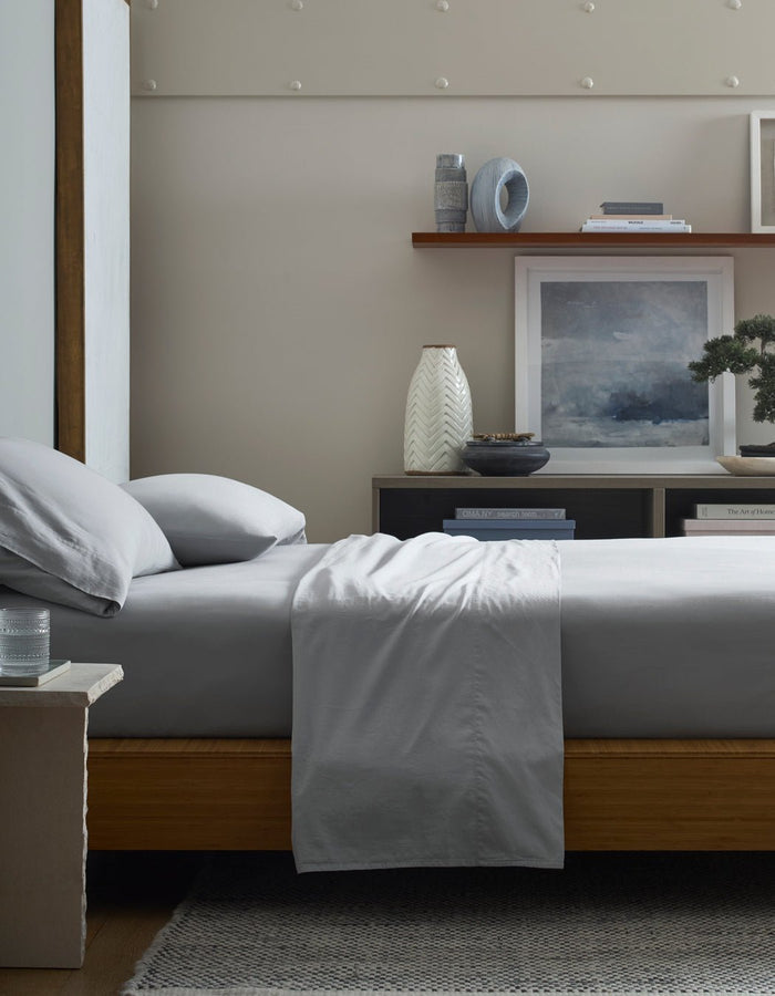 A bed with Cozy Earth Soft-Wash Cotton Fitted Sheet in light gray is styled beside a small table with a glass of water; behind, shelves hold vases, books, and framed art in a modern minimalist bedroom.|Color:Shore