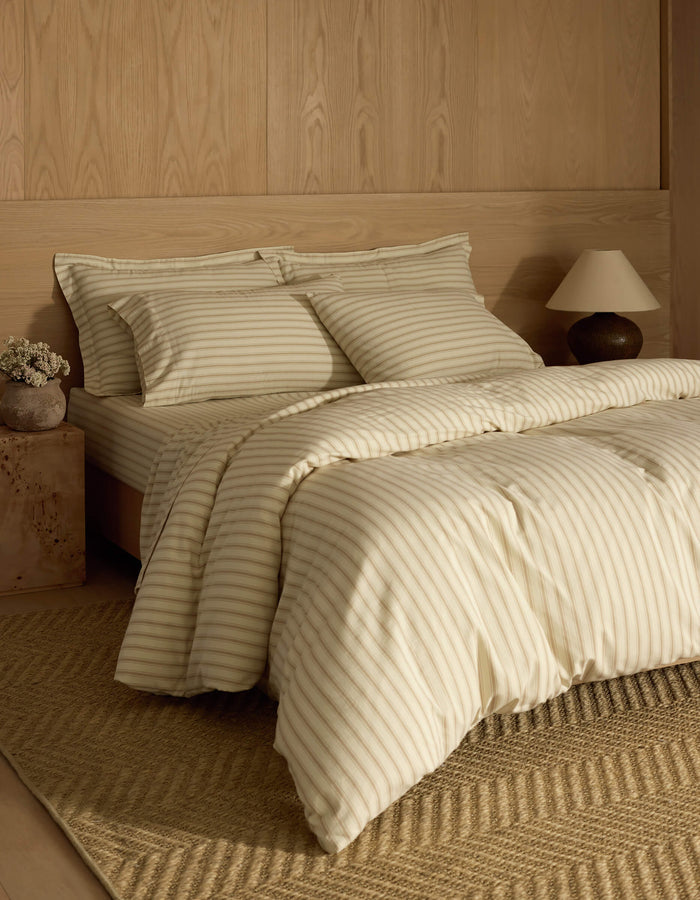 A neatly made bed with Cozy Earth's Soft-Wash Cotton Duvet Cover, matching pillowcases, and a wooden headboard sits against a light wood-paneled wall. A tan woven rug, side table with lamp, and vase of flowers complete the look.|Color:Coconut and Sandalwood Stripe