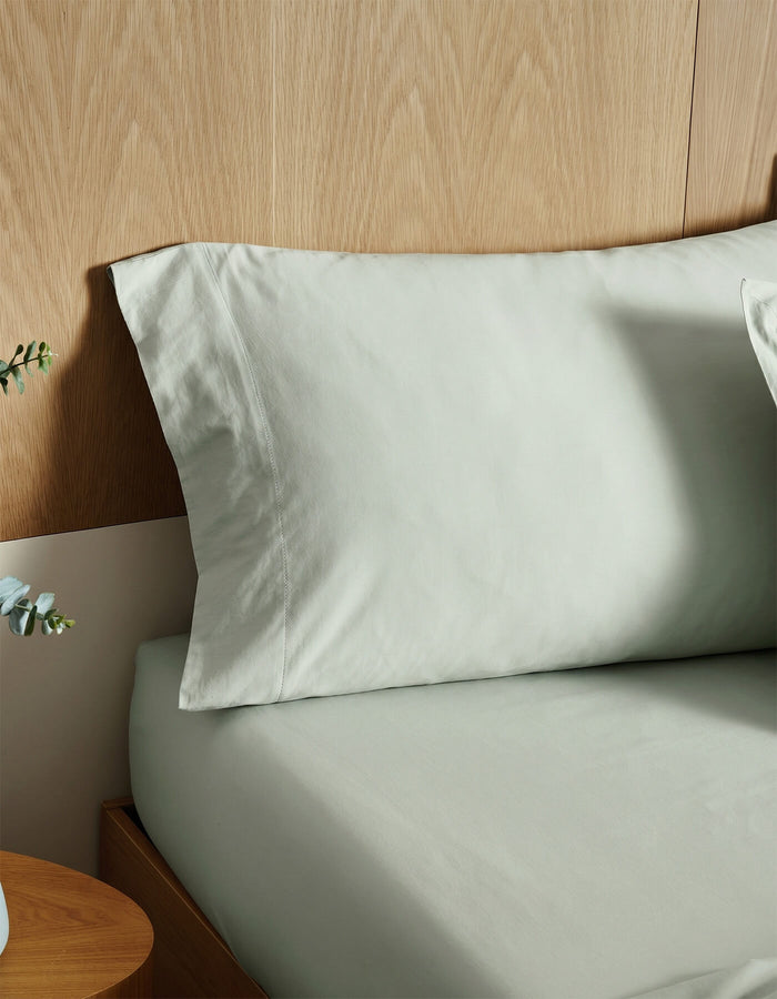 A bed with Cozy Earth's Soft-Wash Cotton Pillowcases in light gray is styled against a light wooden headboard, beside a small wooden nightstand topped with a plant.|Color:Laurel