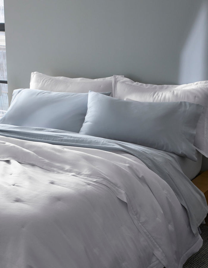 A bed near a window features Cozy Earth Soft-Wash Cotton Pillowcases in light gray and white, with matching bedding and two large pillows, all set against a pale gray wall.|Color:Shore