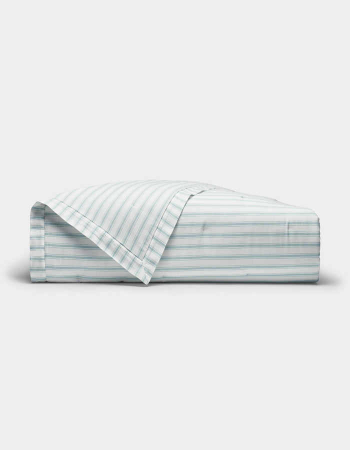 The Cozy Earth Soft-Wash Cotton Quilt is shown neatly folded with one corner turned back, revealing its light color and thin horizontal blue and gray stripes against a plain white background.|Color:River and Sea Salt Stripe