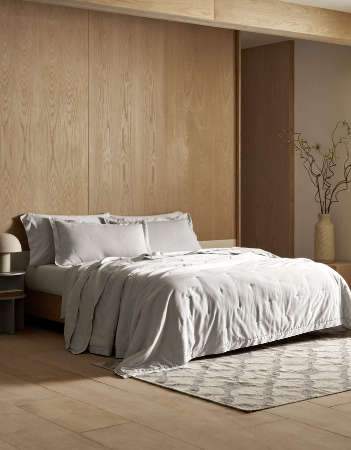 A modern bedroom features light wood-paneled walls, a neatly made bed with the Cozy Earth Soft-Wash Cotton Quilt in light gray, a bedside table with a lamp, a patterned rug, and a tall vase with branches in the corner.|Color:Dew