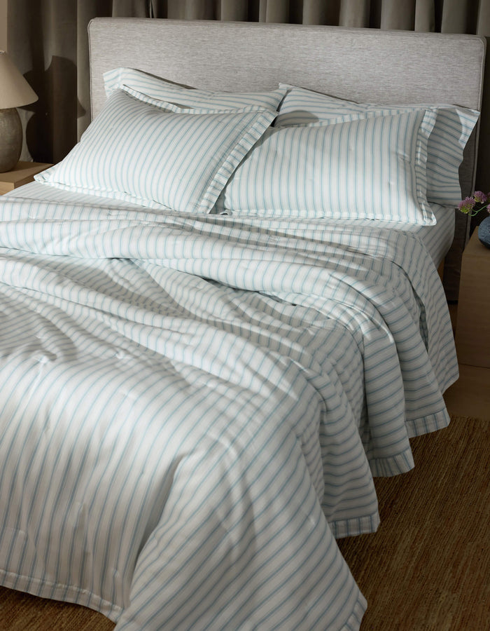A neatly made bed features Cozy Earth's Soft-Wash Cotton Quilted Shams, with blue and white stripes, two pillows, and smooth, inviting bedding in a softly lit room with a light headboard, nightstand, and lamp.|Color:River and Sea Salt Stripe