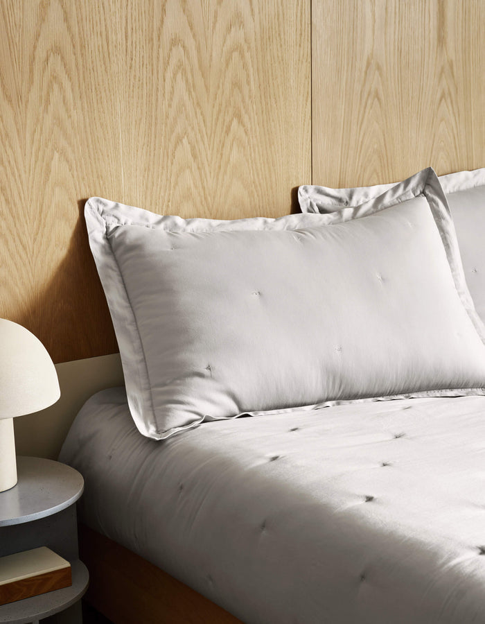 A neatly made bed features the Cozy Earth Soft-Wash Cotton Quilt in light gray with a matching pillow, set against a light wood-paneled wall. On the left, a round bedside table with a white lamp is partially visible.|Color:Dew
