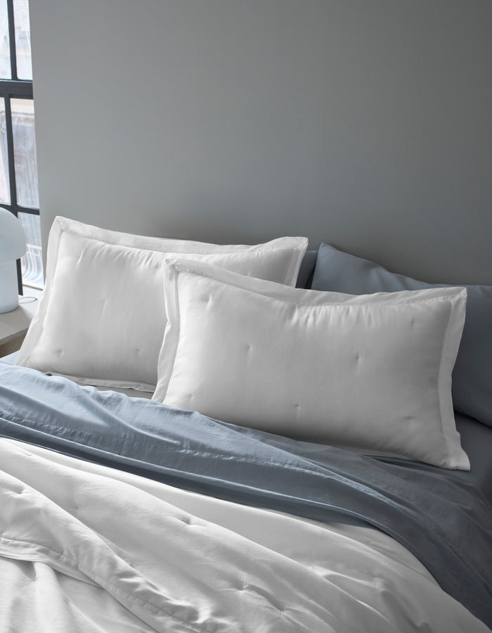 Two Cozy Earth Soft-Wash Cotton Quilted Shams are propped against a gray headboard on a bed with white sheets and a light blue-gray blanket in a softly lit room.|Color:White