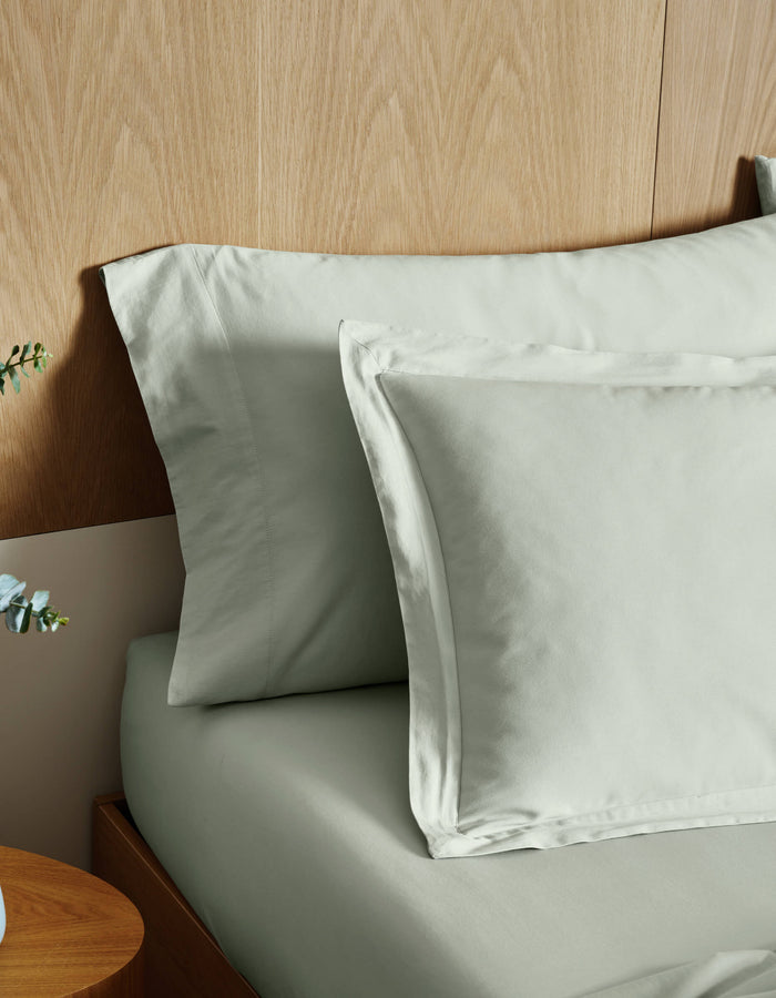 Cozy Earth Soft-Wash Cotton Shams in light gray are neatly arranged on a wooden bed with a wood-paneled headboard; part of a wooden nightstand and a small plant appear in the foreground.|Color:Laurel