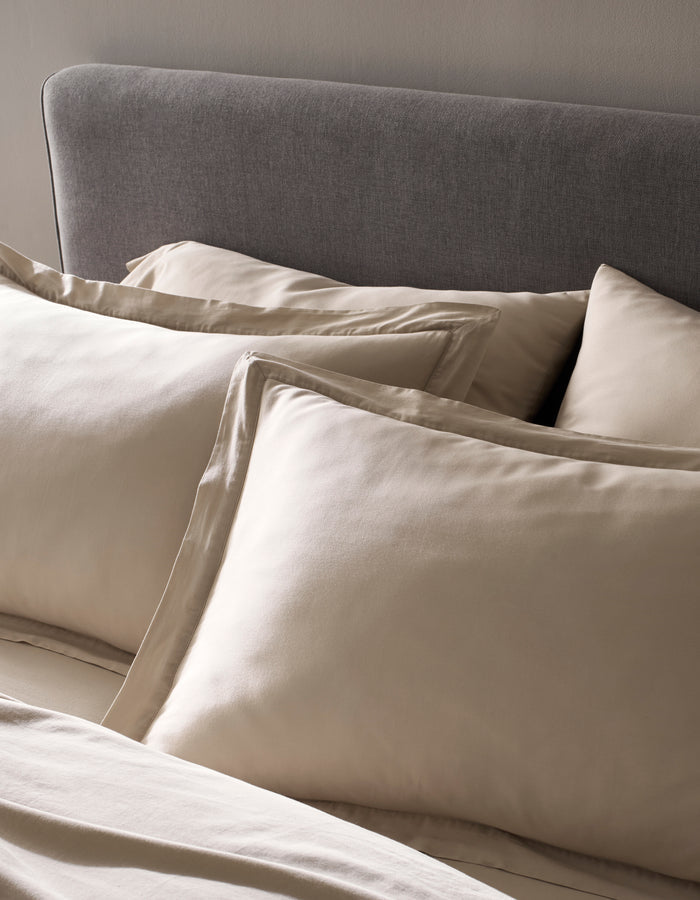 A close-up of a neatly made bed showcases Cozy Earth's Soft-Wash Cotton Shams in beige, paired with matching sheets and a gray upholstered headboard against a neutral wall. The fabric looks exceptionally smooth and soft.|Color:Driftwood