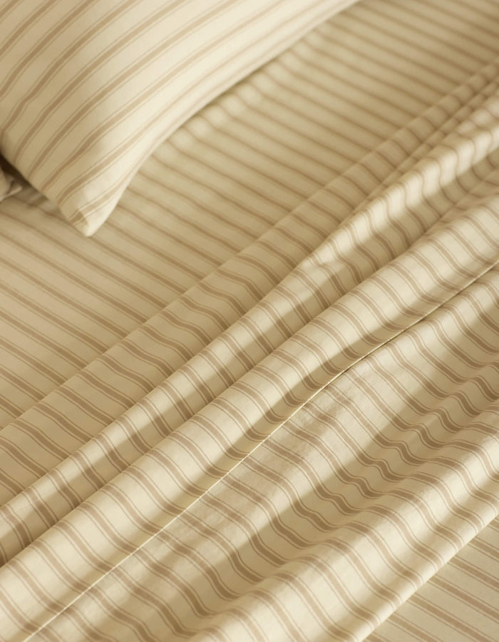 Close-up of a neatly made bed featuring the Cozy Earth Soft-Wash Cotton Sheet Set in beige and cream stripes. The smooth fabric and evenly spaced stripes create a calm, cozy atmosphere.|Color:Coconut and Sandalwood Stripe